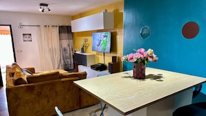 Ein gemütlicher Wohnbereich mit Sofa, kleinem Esstisch, Fernseher und Blumen in einer Vase.