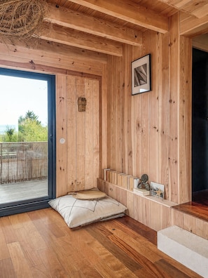 A wooden room with a large window, a cushioned bench, a shelf with decorative items, and a framed picture on the wall.