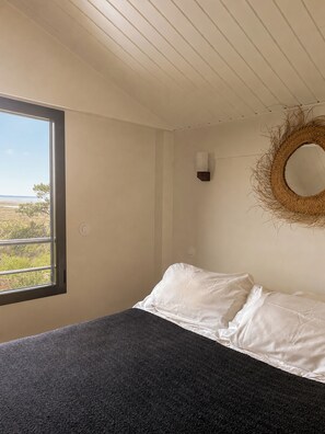 A bedroom with a bed, a window with a view, a wall-mounted light, and a woven wall decoration.
