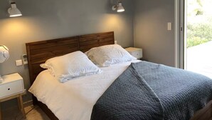 Une chambre à coucher avec une tête de lit en bois, du linge de lit blanc, une couverture matelassée grise, des lampes de chevet et une fenêtre donnant sur un paysage verdoyant.