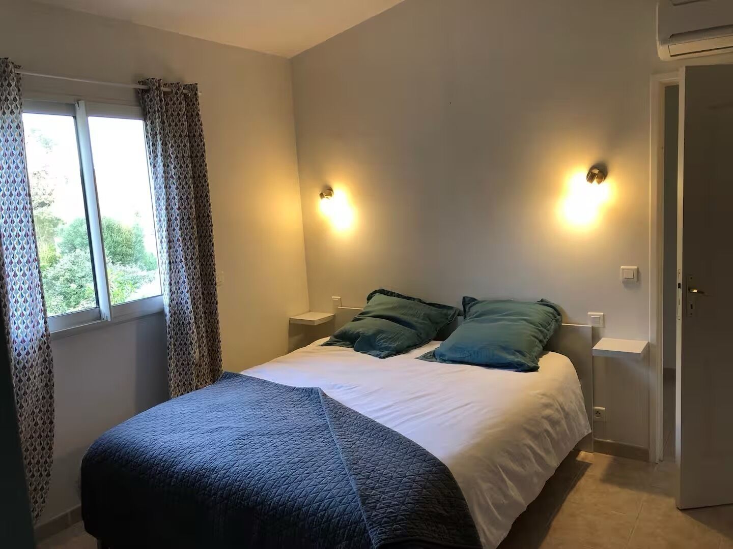 Un dormitorio con cama, dos mesitas de noche, una ventana con cortinas e iluminación en la pared.