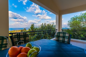 A balcony with a table and chairs, a bowl of fruit, and a scenic view of the sea and mountains. - Maritsa Villa (Neo Chorio)