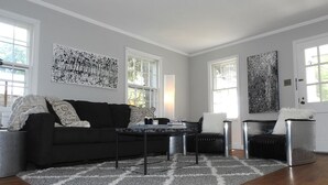 En moderne stue med en sort sofa, et sofabord og et tæppe. Der er to vinduer og en dør.
