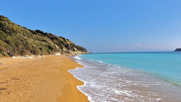 Une plage avec une eau d’un bleu limpide, un rivage sablonneux et un paysage vallonné en arrière-plan.