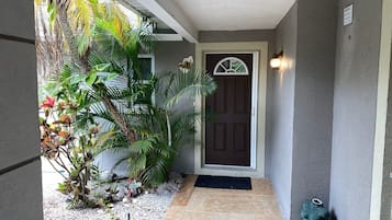 Un porche cubierto con una puerta oscura, un pequeño jardín con plantas tropicales y un suelo de baldosas.