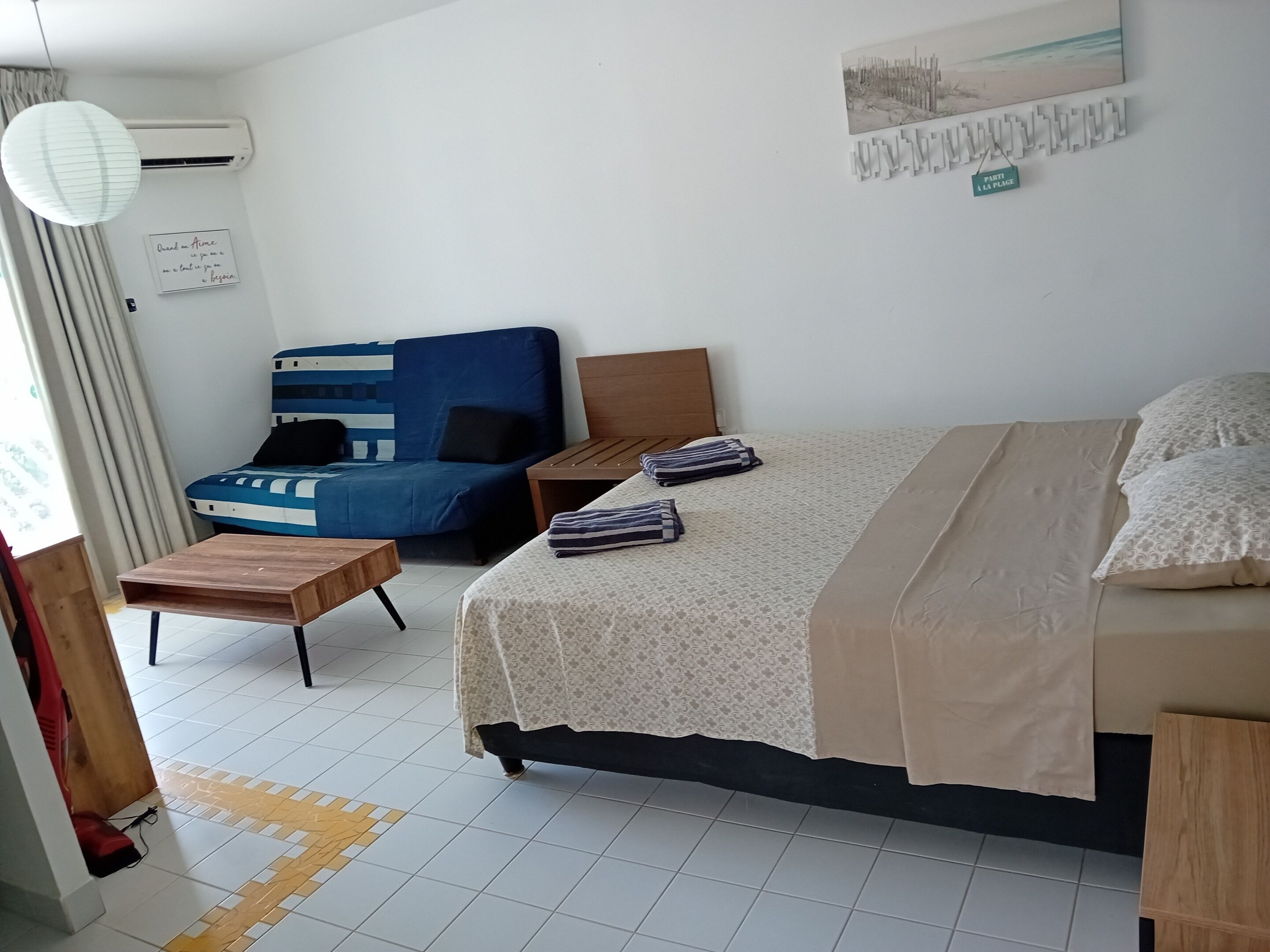 A hotel room with a bed, a sofa, a small table, and a wall-mounted air conditioner.
