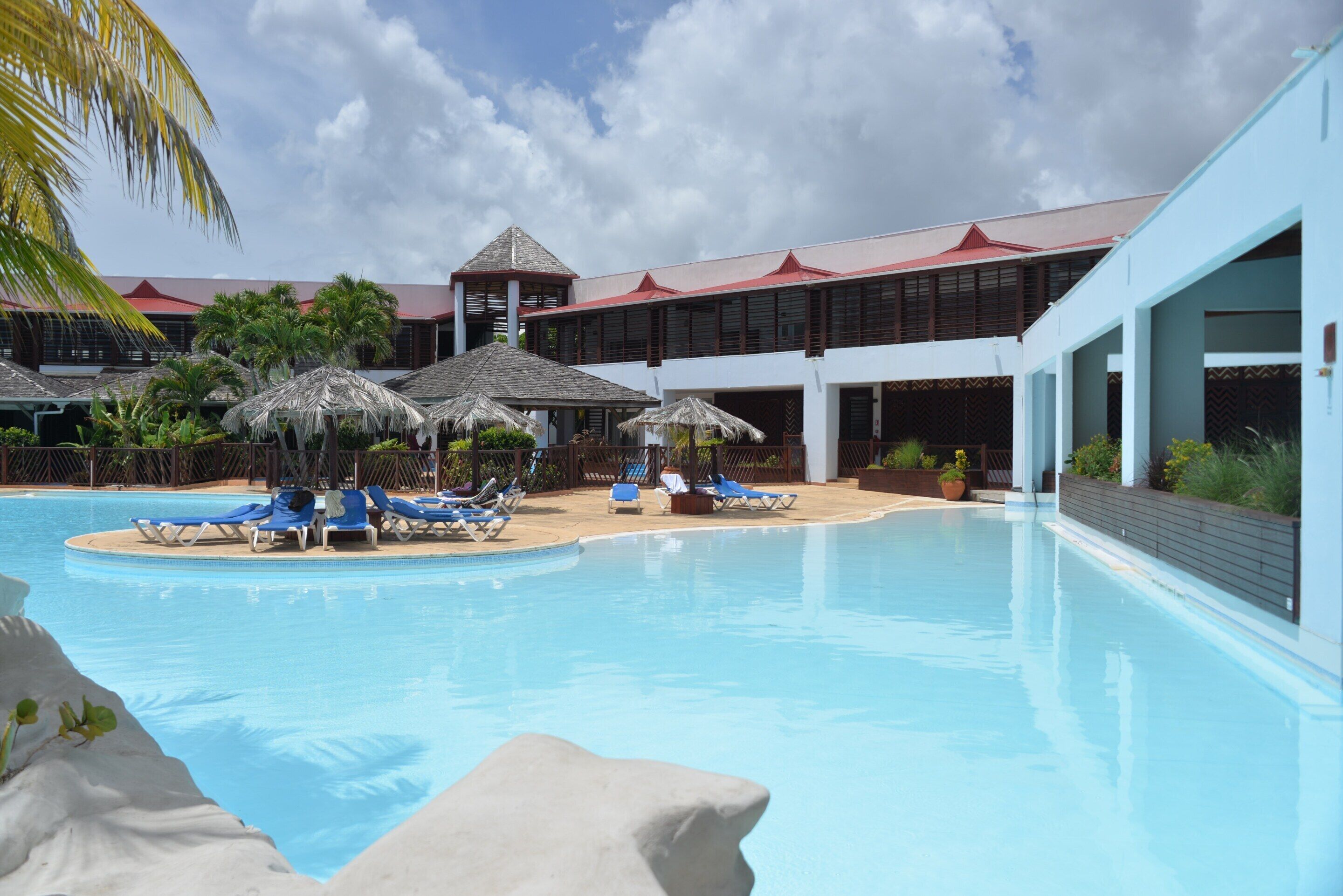 Une piscine entourée de chaises longues et de parasols, le tout bordé par un bâtiment doté d’un toit rouge et d’une façade blanche.