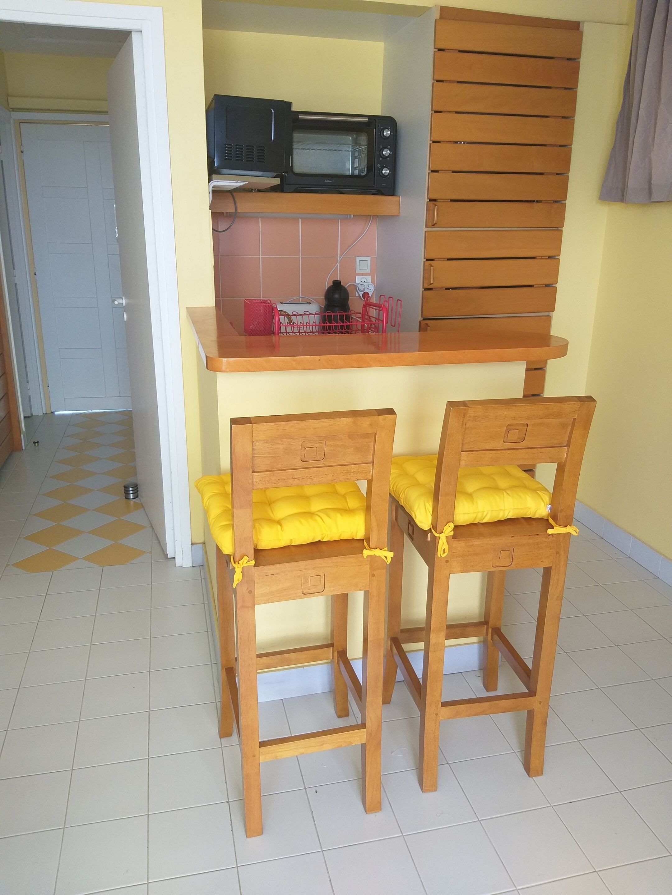 Eine kleine Küchenzeile mit einer Mikrowelle und einem Toaster, zwei Holzstühle mit gelben Kissen und ein gefliestes Badezimmer.