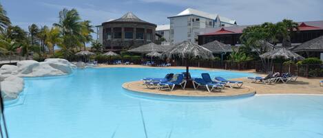 A pool with lounge chairs, palm trees, and a building with a red roof in the background.