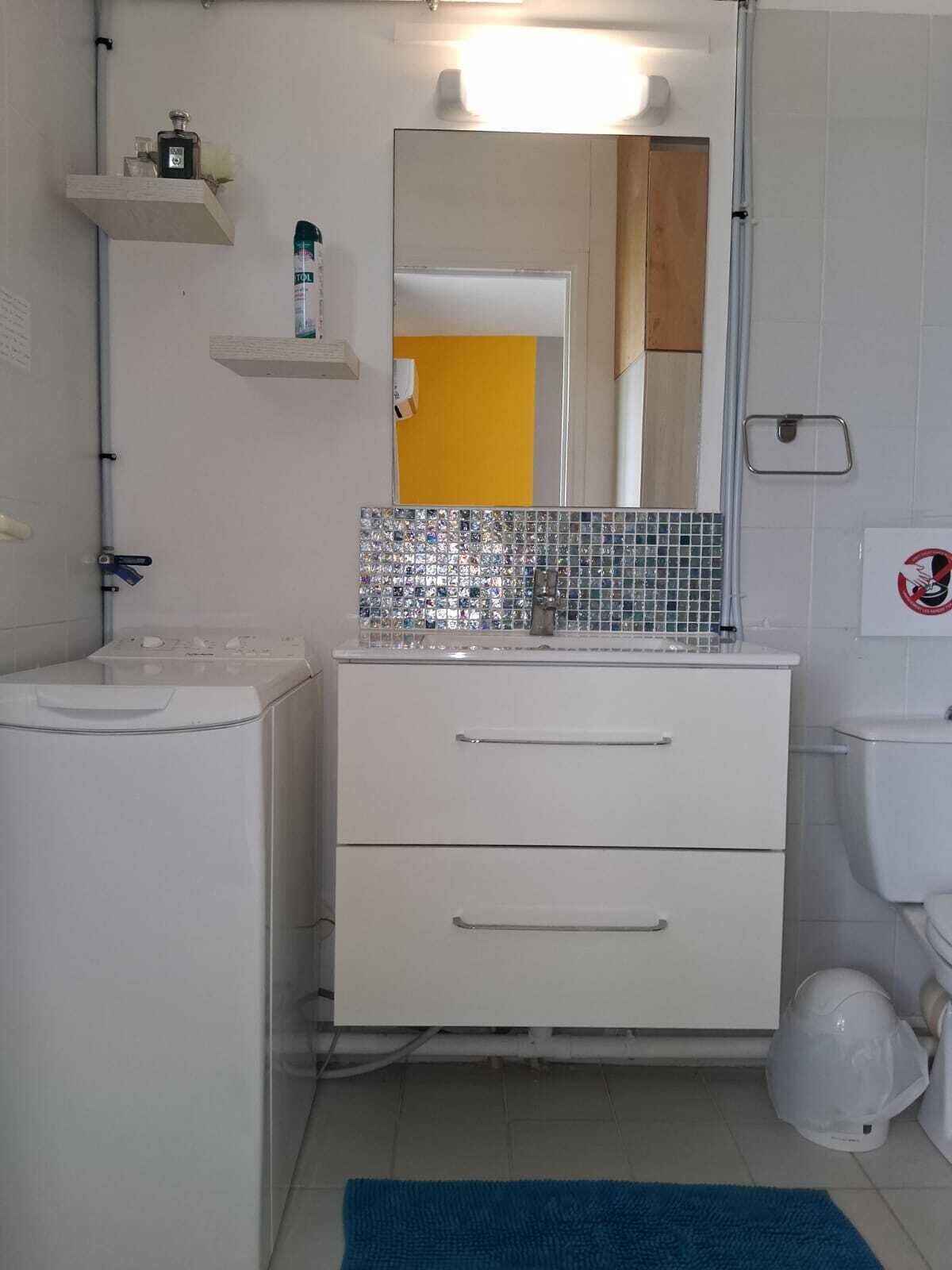 Une salle de bain avec un lavabo blanc, un miroir et des toilettes. Il y a deux tiroirs sous le lavabo et un tapis bleu sur le sol.