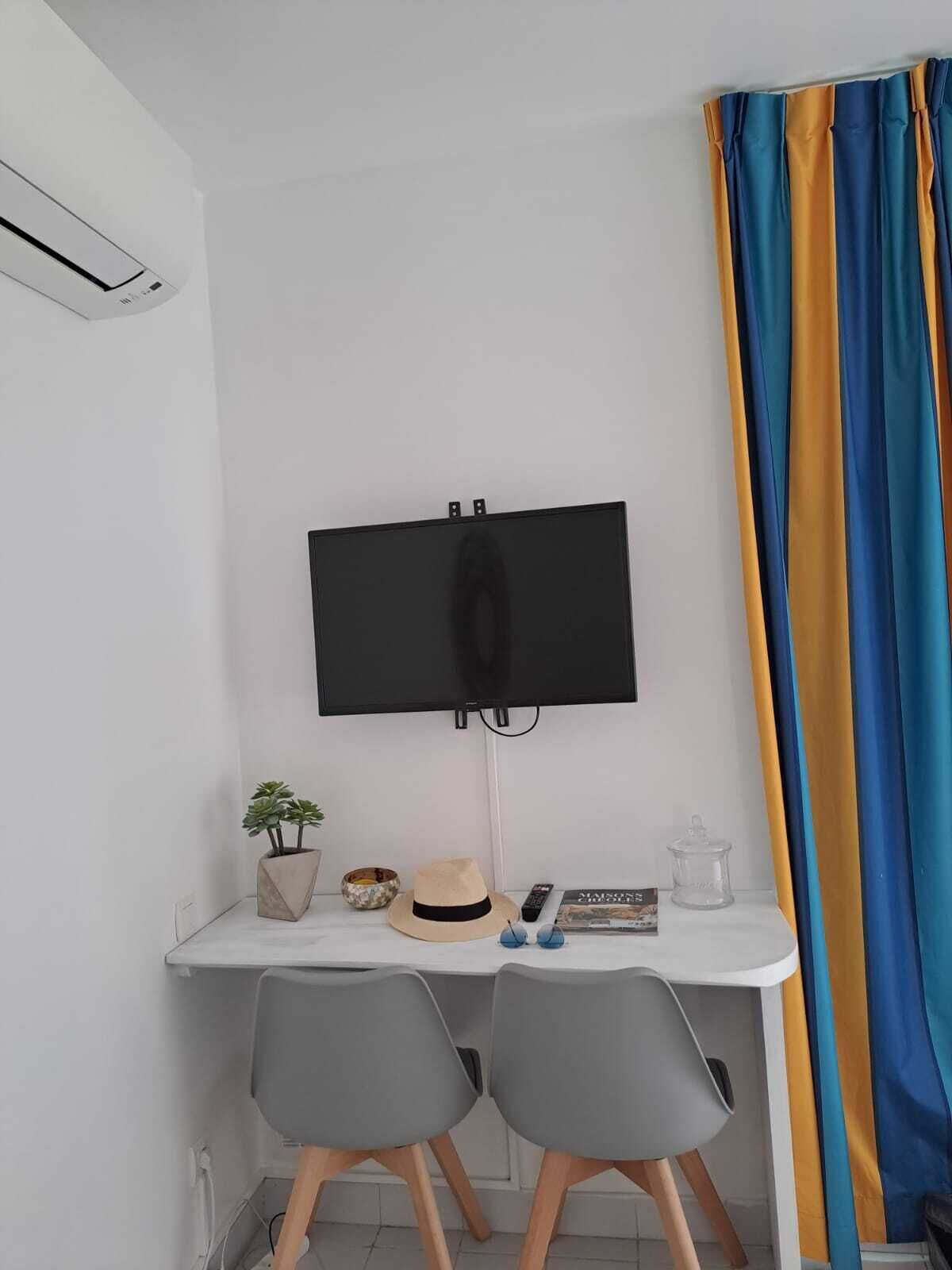 Una habitación pequeña y moderna con un escritorio blanco, dos sillas grises, un televisor de pantalla plana montado, una planta en maceta, un sombrero, un control remoto y una cortina a rayas.