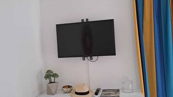 Una habitación pequeña y moderna con un escritorio blanco, dos sillas grises, un televisor de pantalla plana montado, una planta en maceta, un sombrero, un control remoto y una cortina a rayas.