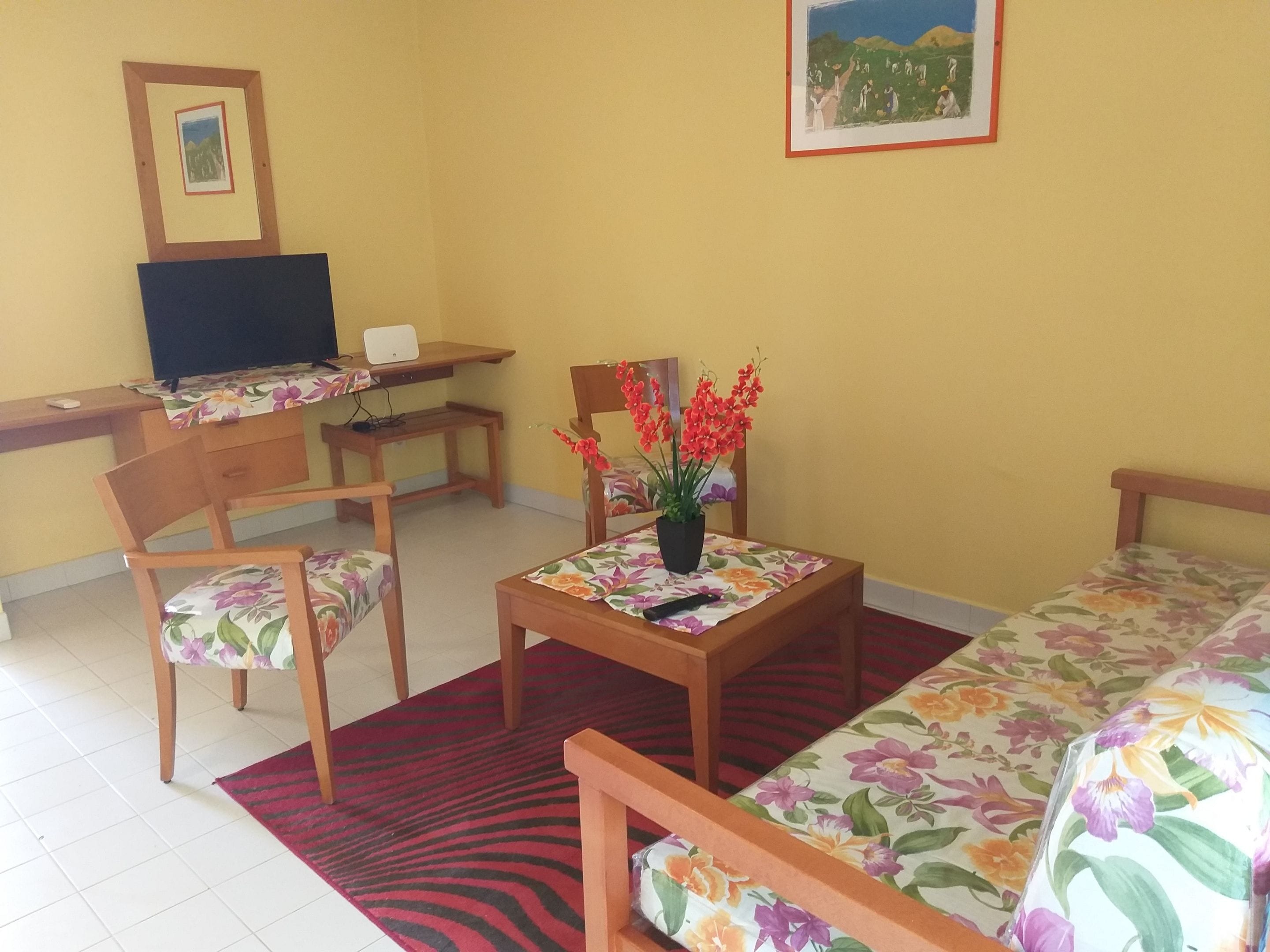 A room with a bed, a desk, a chair, a small table with a potted plant, and a red rug.