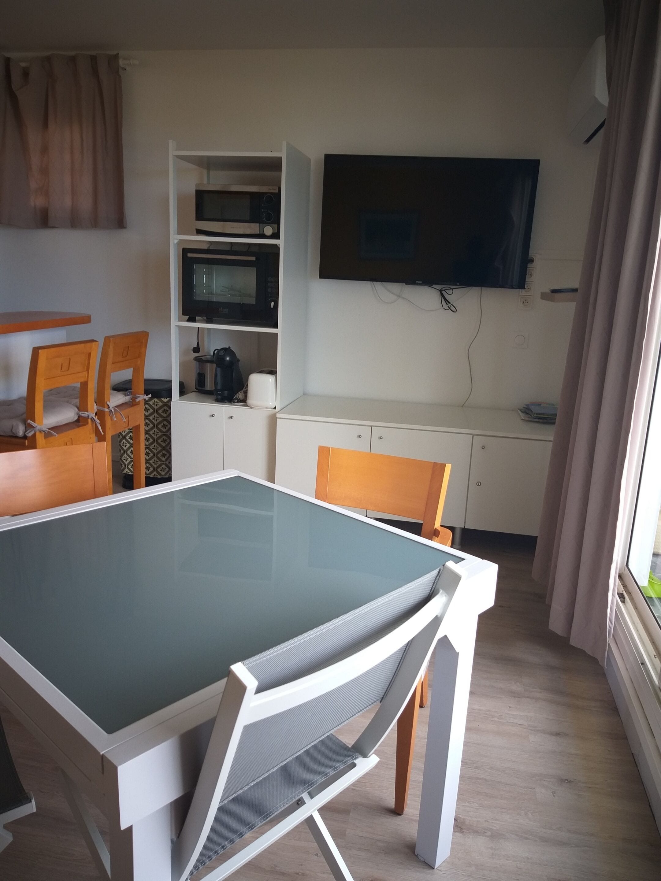 Un espace de vie compact comprenant une table à manger avec un plateau en verre, deux chaises en bois, un four à micro-ondes, un téléviseur et un petit coin cuisine.