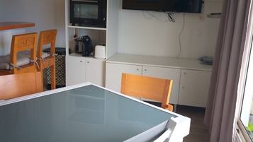 Un espace de vie compact comprenant une table à manger avec un plateau en verre, deux chaises en bois, un four à micro-ondes, un téléviseur et un petit coin cuisine.