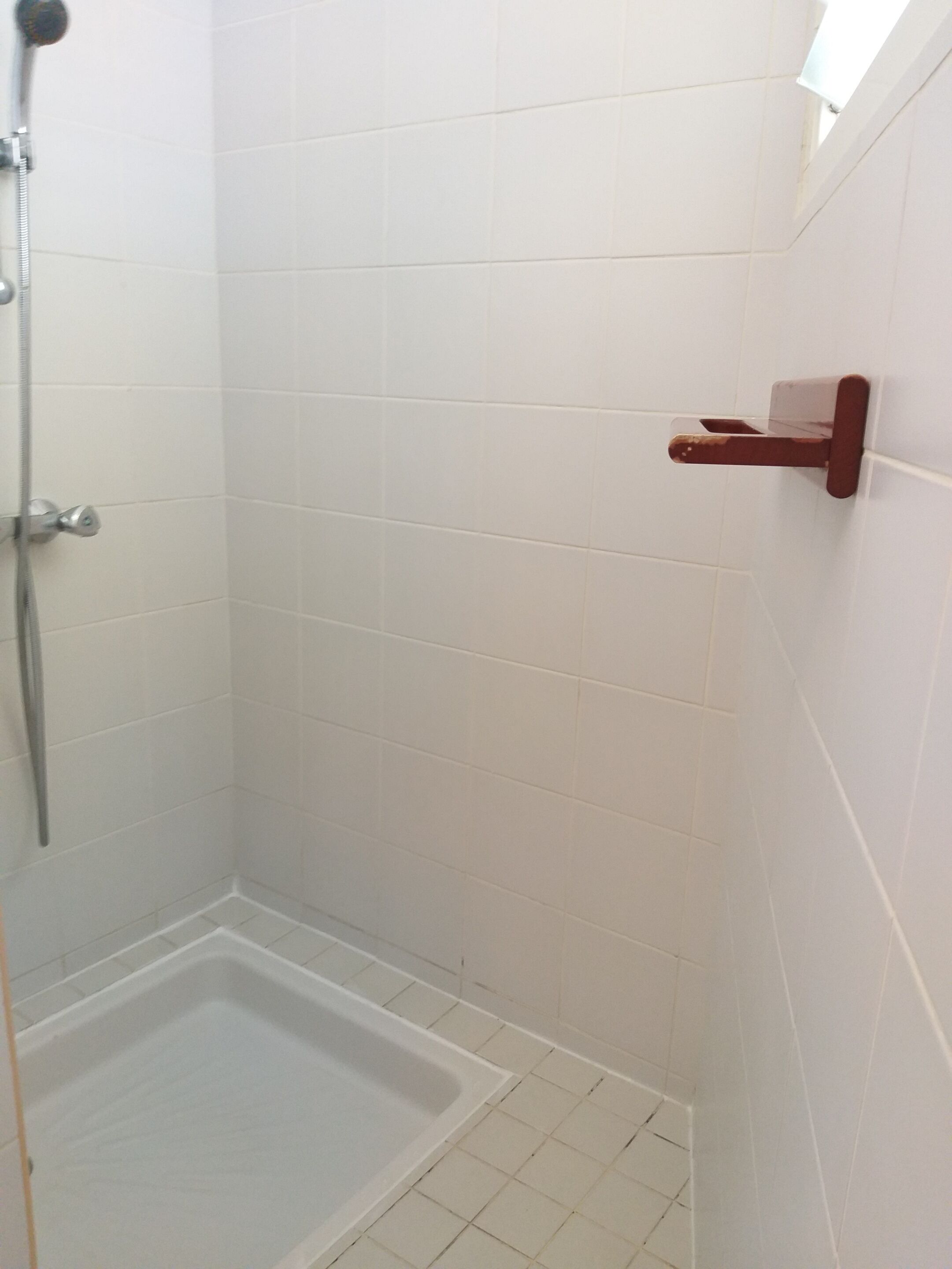 A small, enclosed shower area with white tiled walls and floor, a showerhead, and a wooden towel rack.