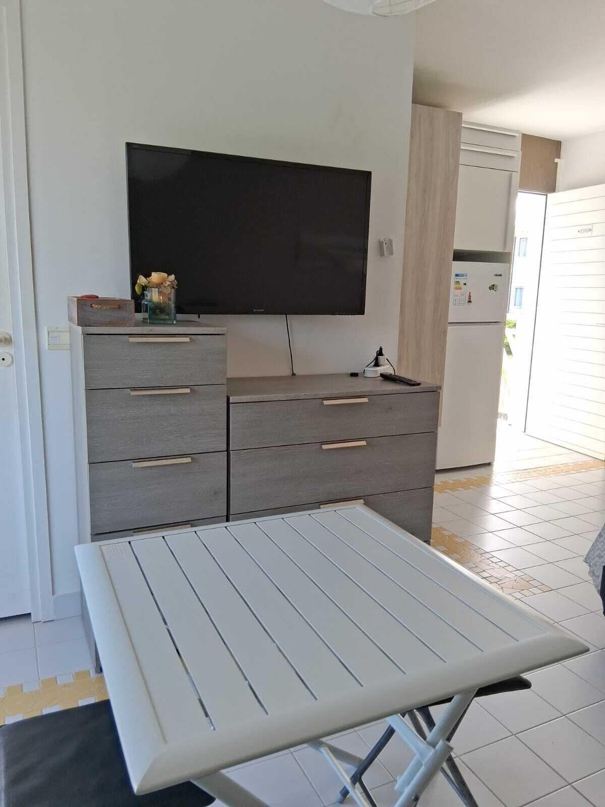 A modern kitchen with a flat-screen TV, grey cabinets, and a white table with a metal frame.