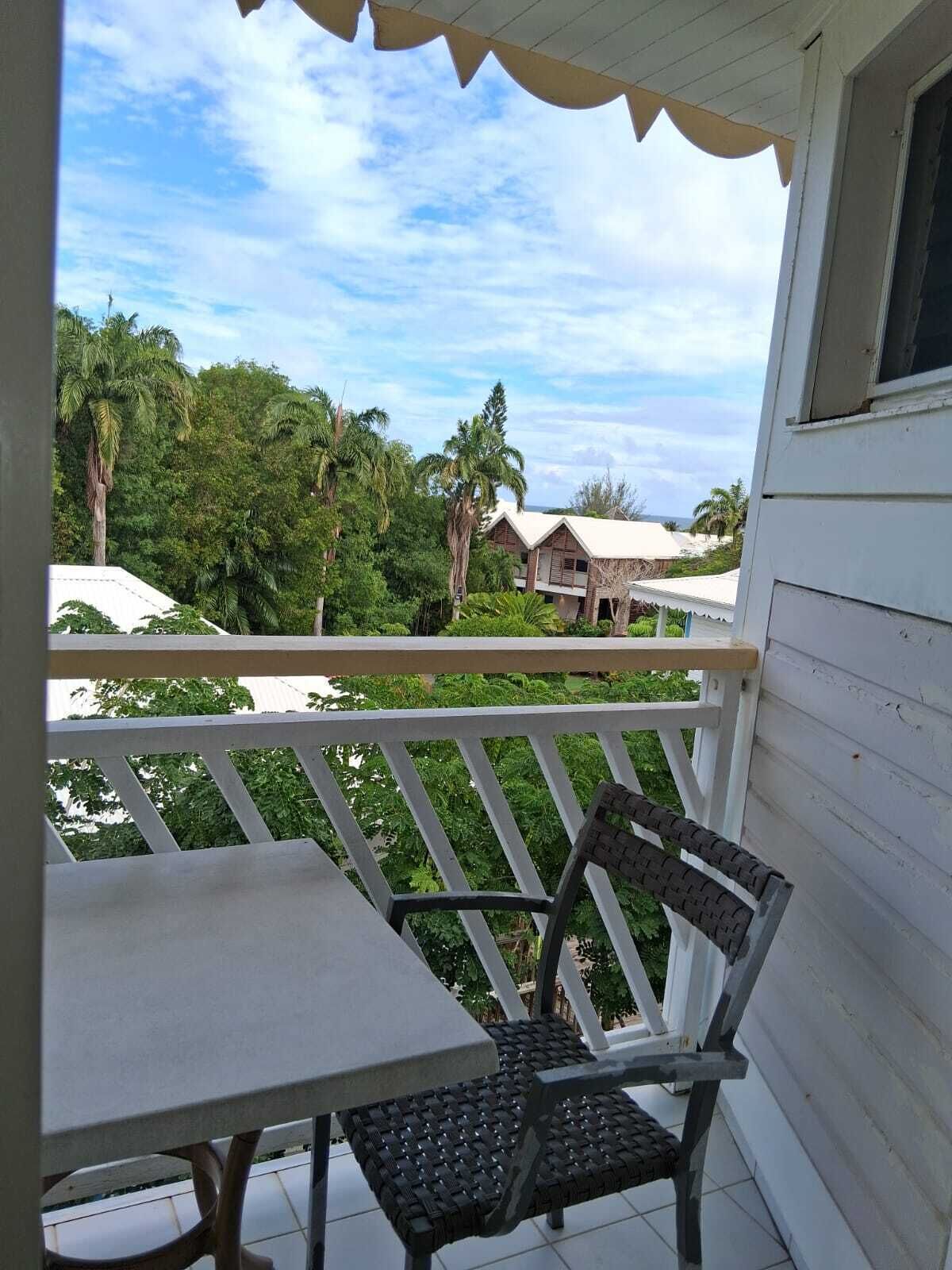 Un balcon avec une table et une chaise, donnant sur un paysage verdoyant et luxuriant et sur un bâtiment doté d’un toit blanc.