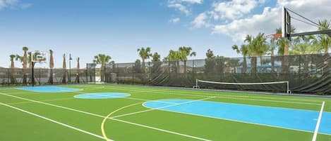 An outdoor basketball court with a net, surrounded by a fence, and palm trees in the background.