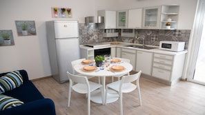A modern kitchen with white cabinets, a built-in oven, and a microwave. There is a dining table with chairs and a small potted plant.