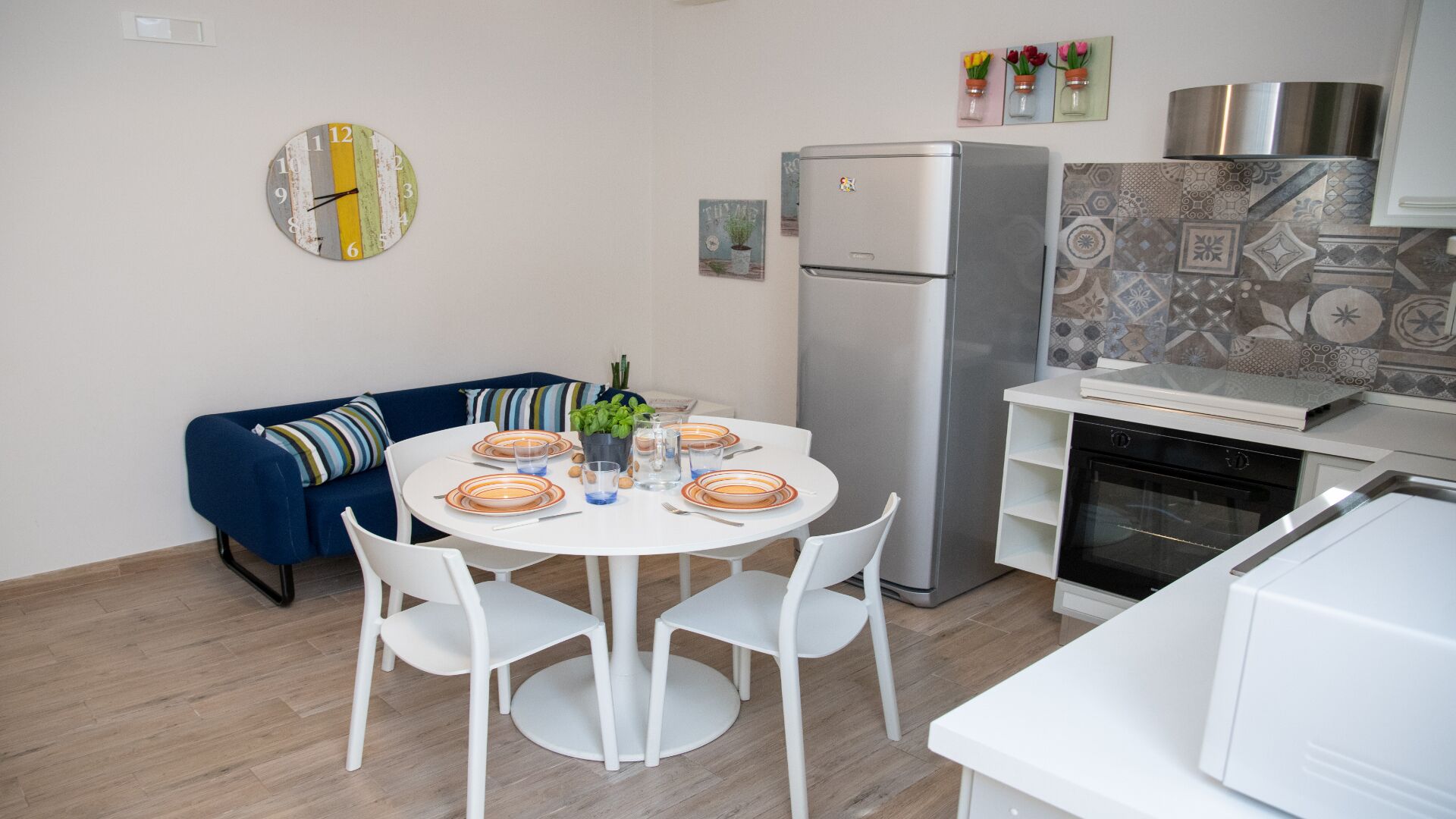 A compact kitchen with a refrigerator, a dining table set with plates and glasses, and a blue sofa with striped cushions.