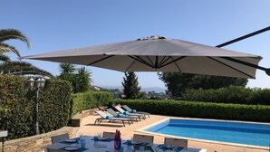 Un área de comedor junto a la piscina con una mesa preparada para cuatro personas, una sombrilla grande y sillones de descanso.