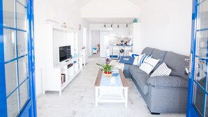En rummelig opholdsstue med en grå sofa, et sofabord og et fjernsyn. I baggrunden er der et køkken med hvide skabe og en blå bordplade.