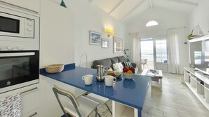 Et moderne køkken med en blå bordplade, et spiseområde med en grå sofa, og havudsigt gennem skydedøren.