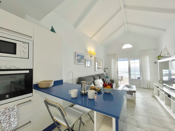 A modern kitchen with a blue countertop, a dining area with a gray sofa, and a view of the ocean through the sliding glass door.