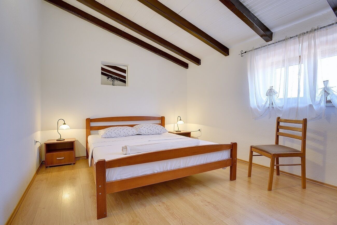 A bedroom with a wooden bed, bedside tables with lamps, a wooden chair, and a window with sheer curtains.