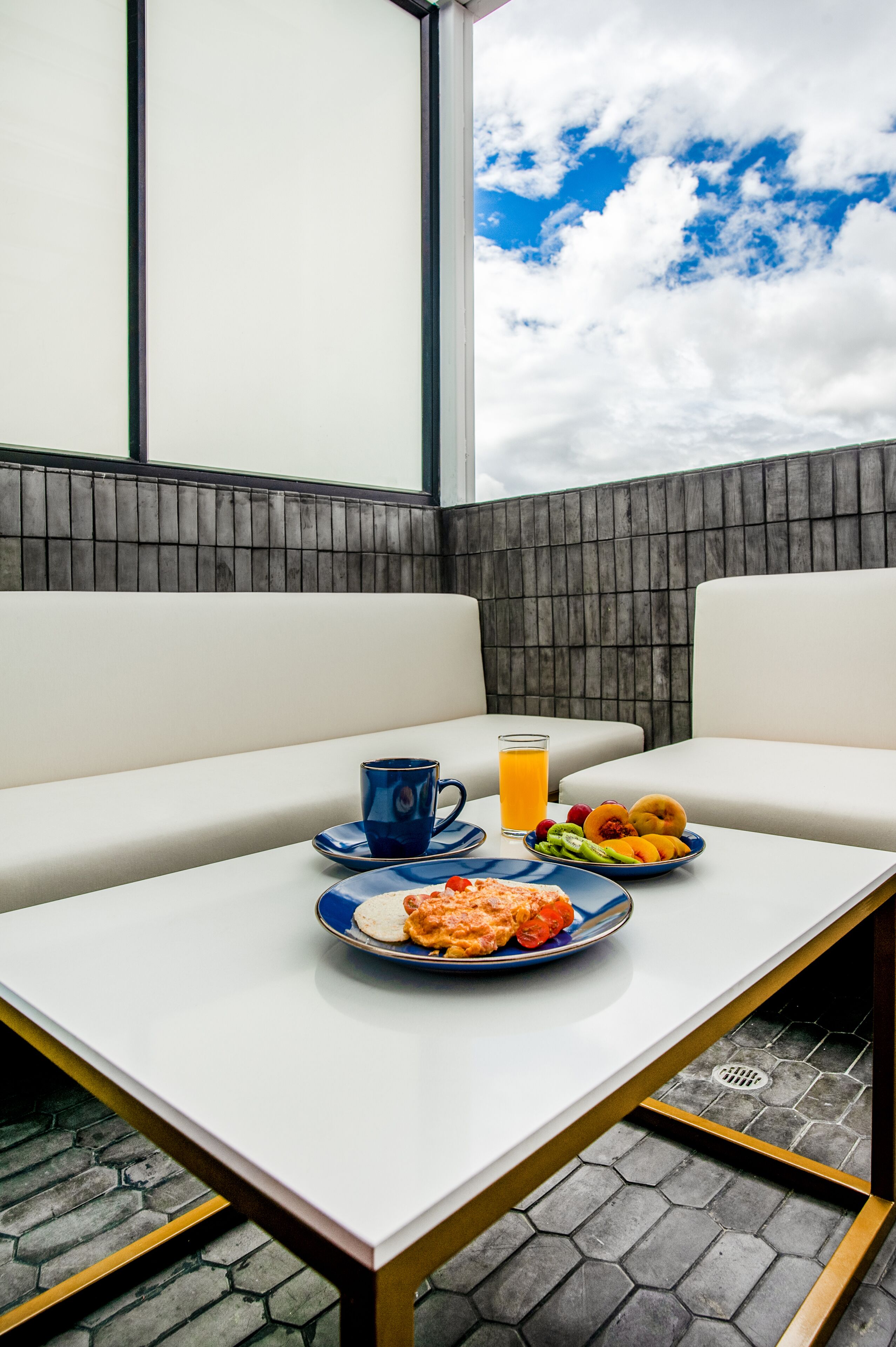 A breakfast setup with a plate of food, a blue mug, and a glass of orange juice on a white table in a room with a view of the sky.
