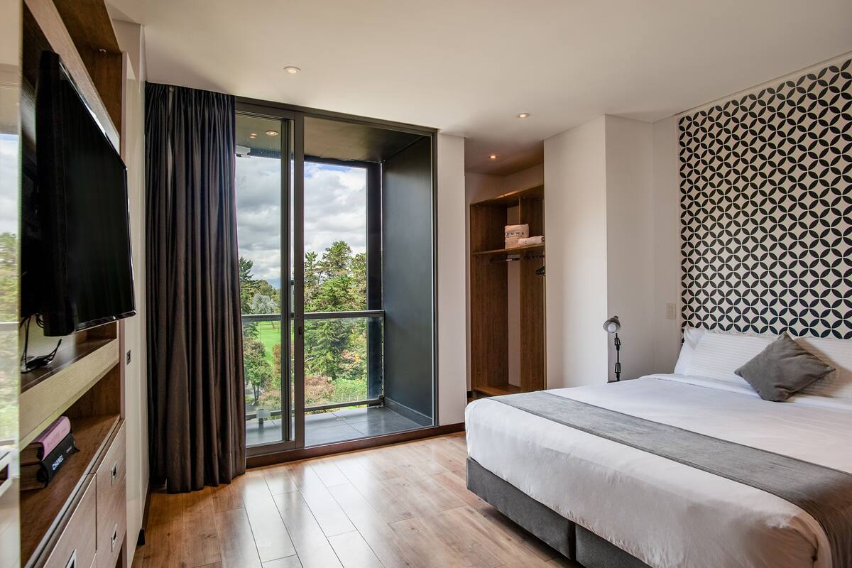 A modern hotel room with a large bed, a TV, a wardrobe, and a view of greenery through a large window.