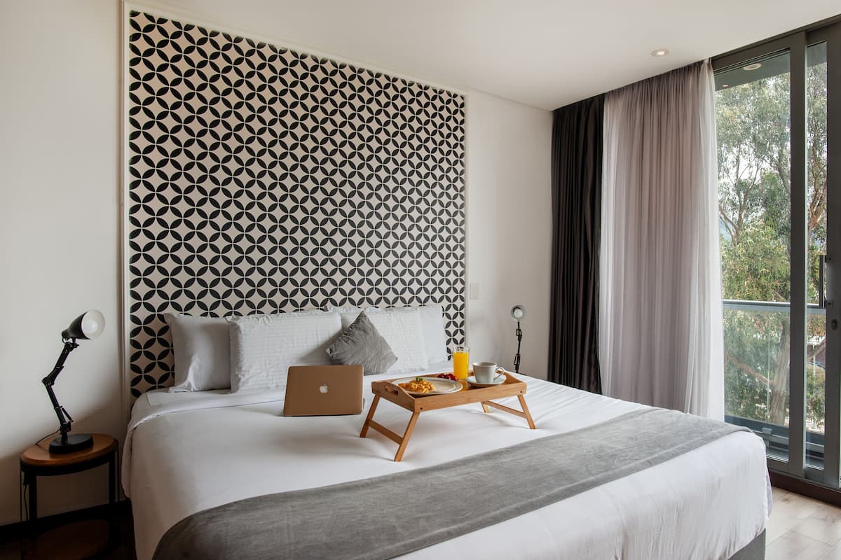 A modern hotel room with a geometric-patterned headboard, a laptop on the bed, a tray with breakfast, and a view of greenery through the window.