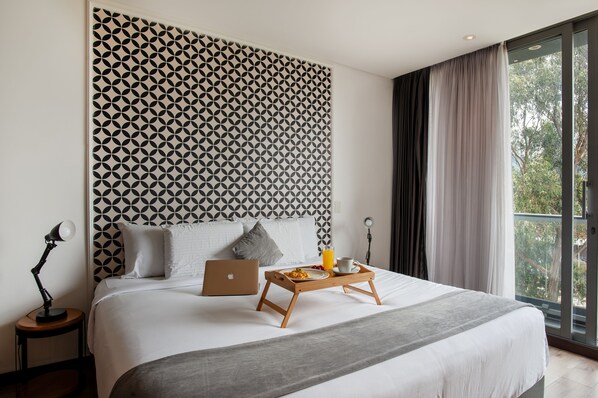 A modern hotel room with a geometric-patterned headboard, a laptop on the bed, a tray with breakfast, and a view of greenery through the window. - Hemma Bogotá Country Luxury Suites Hotel (Bogotá)