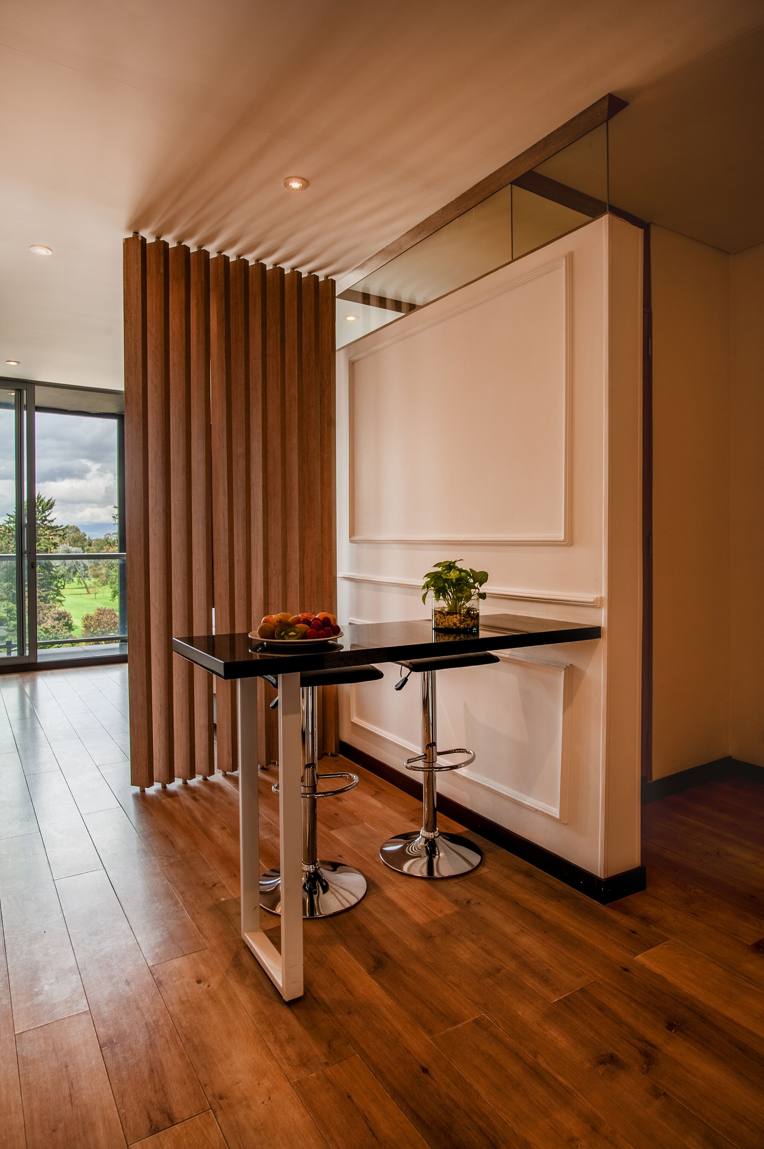Une pièce moderne avec un plancher en bois, une table noire sur laquelle se trouve un saladier rempli de fruits, et une plante en pot.