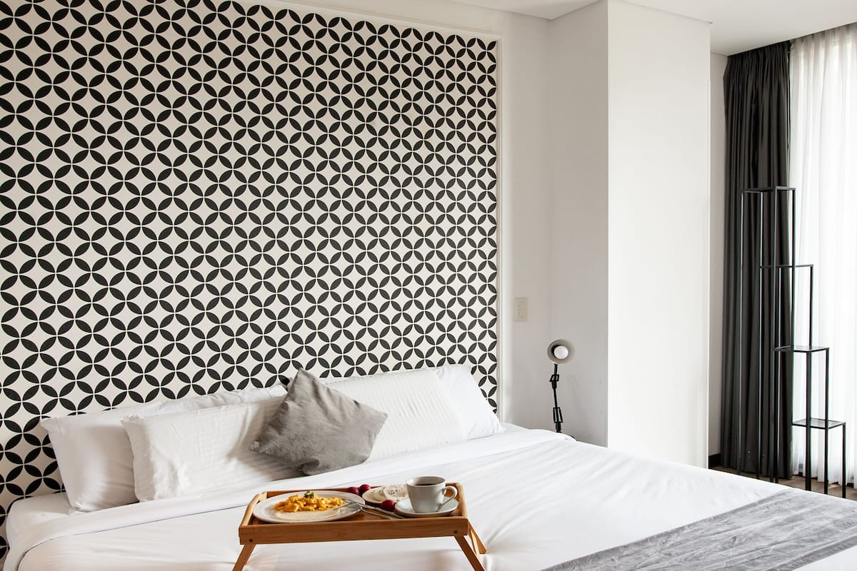 A neatly made bed with a tray of food, a patterned wall, and a window with curtains.