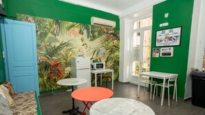 Una habitación con pared verde, puerta azul, una cama, una mesa blanca, una mesa roja, una silla blanca, un cubo de basura negro y un aire acondicionado instalado en la pared.