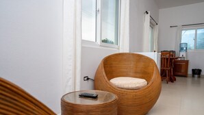 A modern living room with a large wicker chair, a small round table with a remote control, and a dining area with wooden furniture. - "Experience Tranquility at Hibiscus Studio" (Caye Caulker)