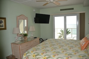 Quarto com cama, penteadeira e TV fixada na parede. Possui varanda com vista para o mar.