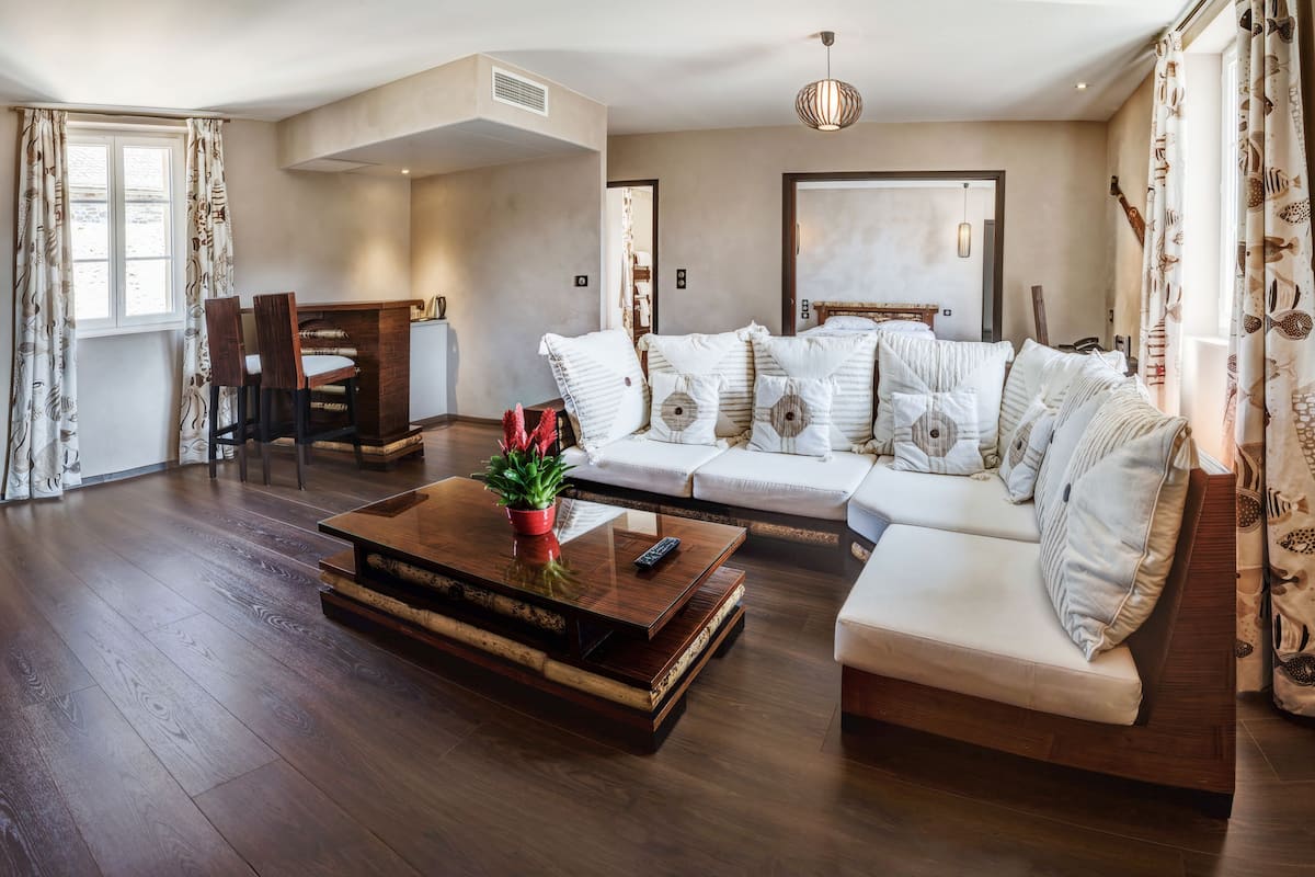 A spacious living room with a wooden floor, a white sofa with patterned cushions, a coffee table with a potted plant, and a dining area with a table and chairs.