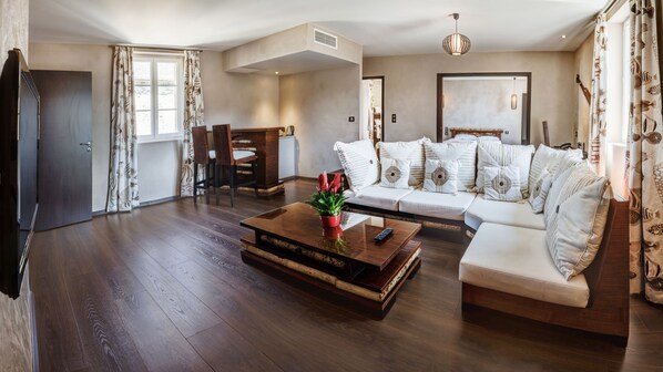 A spacious living room with a wooden floor, a white sofa with patterned cushions, a coffee table with a potted plant, and a dining area with a table and chairs. - Château de la Falque, The Originals Relais (Saint-Geniez-d'Olt-et-d'Aubrac)