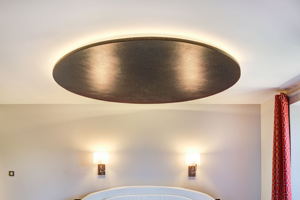 A modern bedroom with a circular bed, two bedside tables, a large circular ceiling light, and a red patterned curtain.