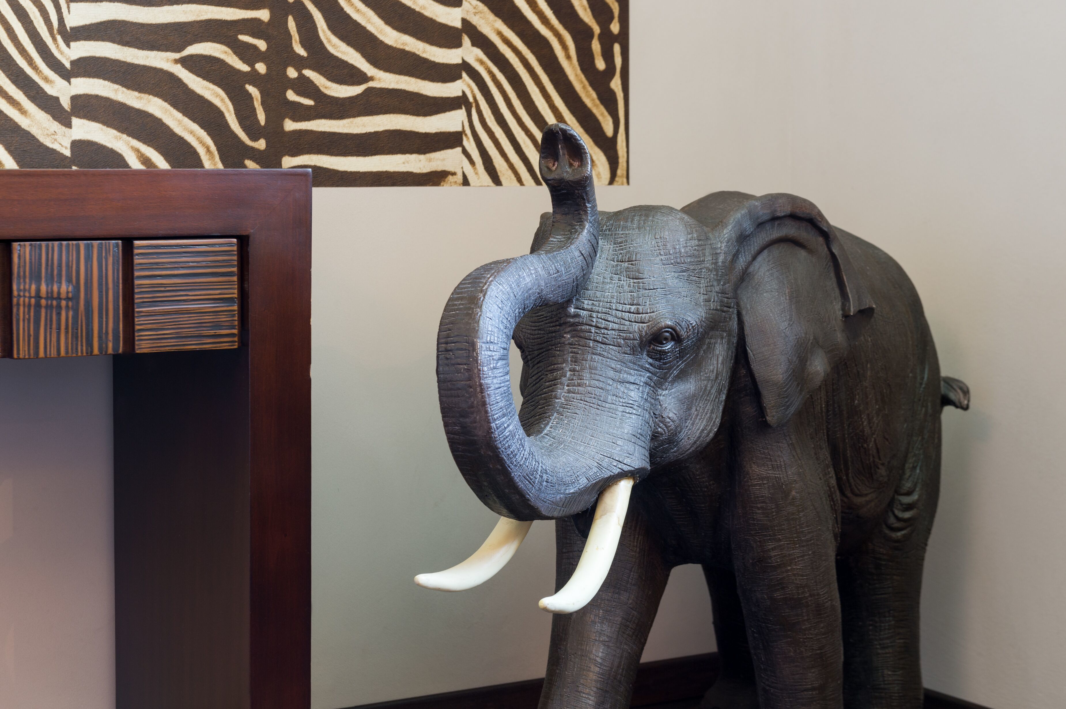 Una escultura de madera de un elefante con colmillos, situada frente a un fondo con estampado de cebra.