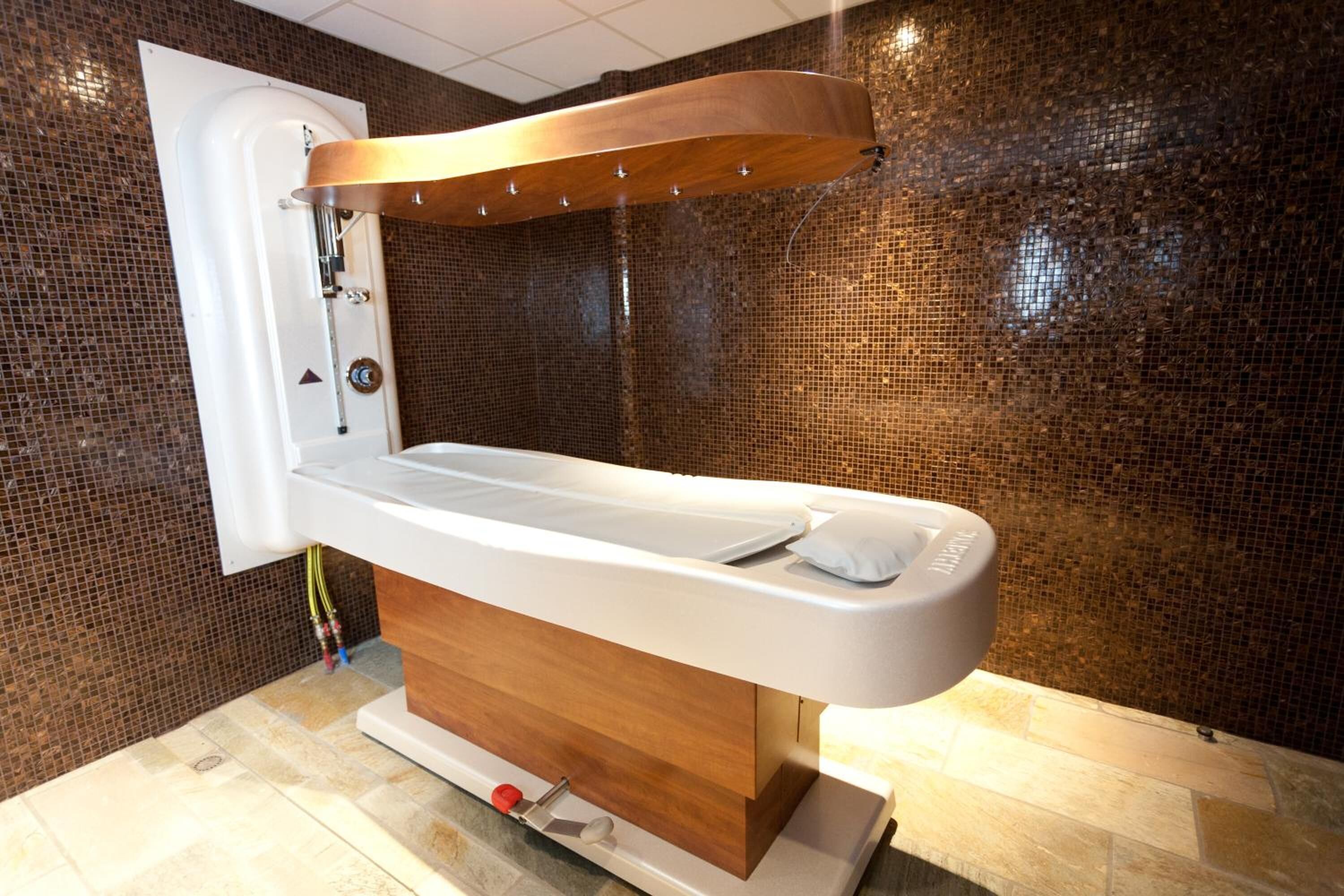 Ein moderner Wellnessraum mit einem Holzmassagetisch, einer an der Wand montierten Duschbrause und einer Wand aus Mosaikfliesen.