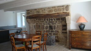A dining area with a stone fireplace, wooden table and chairs, a lamp on a dresser, and a kitchen in the background. - STONE HOUSE RENOVATED WITH TASTE WITH TENNIS IN A HAVEN OF PEACE (Féniers)