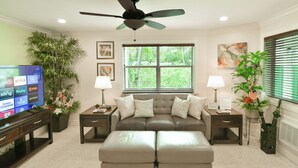 Sala de estar con sofá, reposapiés, televisor y plantas decorativas.