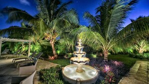 Un espacio exterior nocturno con una fuente escalonada, palmeras y sillones de descanso.