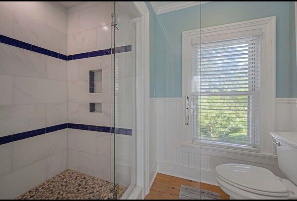 A bathroom with a glass shower enclosure, a window with blinds, and a white toilet.