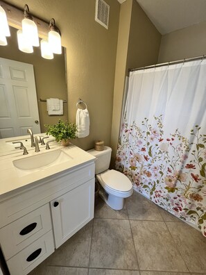 Un baño con lavamanos, tocador y retrete blancos. Hay una cortina de ducha con estampado floral.