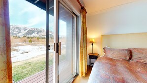 Une chambre à coucher comprenant un lit, une table de chevet, une lampe et une porte-fenêtre coulissante donnant sur un espace extérieur enneigé, avec des montagnes en arrière-plan.